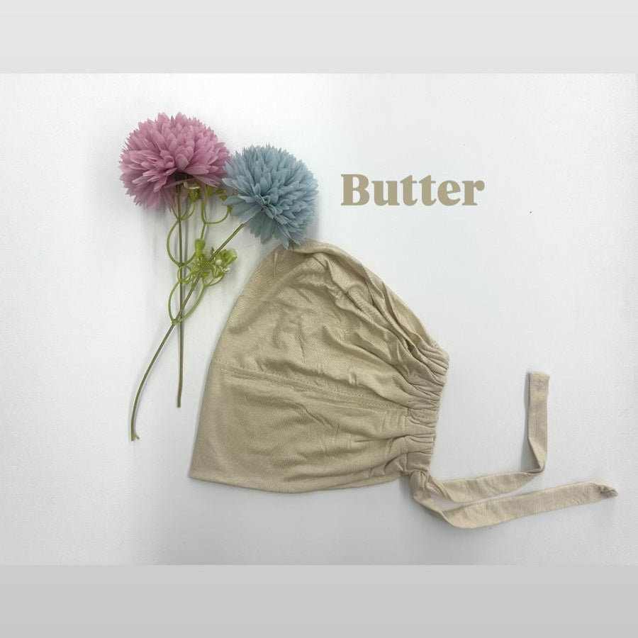 Beige baby hat with a ribbon and two pom-poms on a white background, featuring the brand 'Butter'.